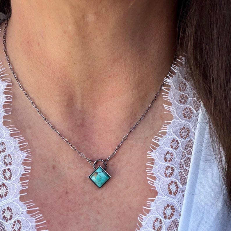 May include: A silver-toned chain necklace with a square turquoise pendant. The pendant is set in a dark metal frame. The necklace is worn against a white lace background.