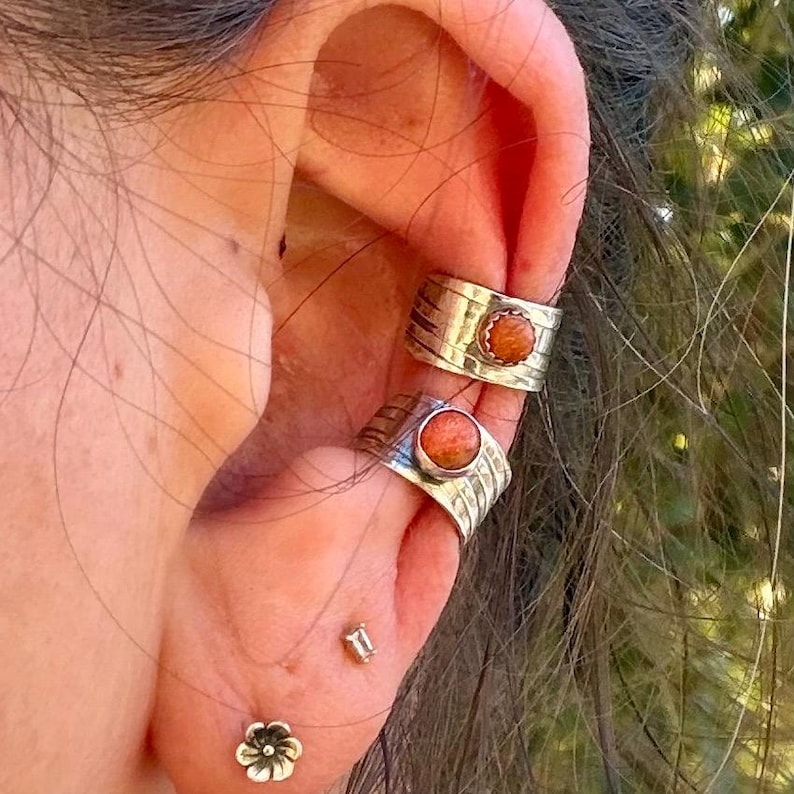 May include: Two silver ear cuffs featuring textured bands and round, orange-brown stone inlays. Below these, a small silver flower-shaped stud earring is worn in the earlobe, accompanied by a tiny silver ball stud earring.