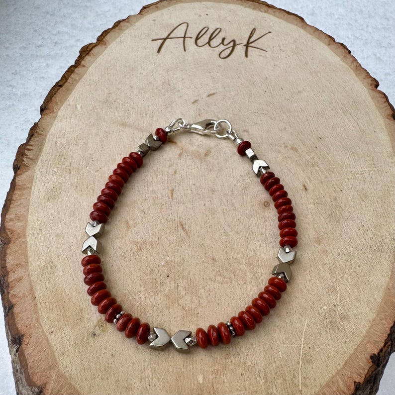 May include: A red beaded bracelet with silver-toned accents and a clasp. The bracelet features small, round red beads interspersed with silver arrow-shaped beads. The clasp is silver and the bracelet is displayed on a wooden surface. The text "Allyk" is visible.