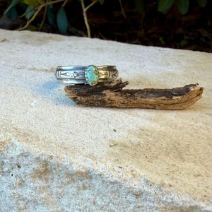 Puede incluir: Un brazalete de plata tipo pu&ntilde;o con una piedra turquesa ovalada engastada en el centro. La banda de metal presenta intrincados patrones grabados a lo largo de su superficie, mostrando un acabado pulido que contrasta con la piedra azul verdosa.