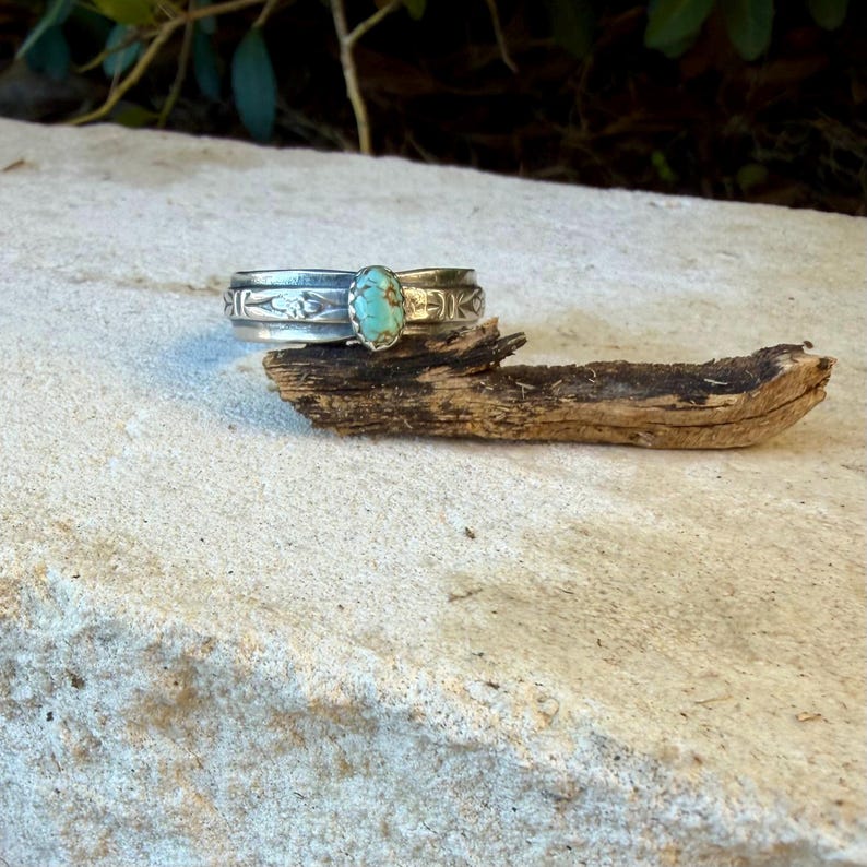 Puede incluir: Un brazalete de plata tipo pu&ntilde;o con una piedra turquesa ovalada engastada en el centro. La banda de metal presenta intrincados patrones grabados a lo largo de su superficie, mostrando un acabado pulido que contrasta con la piedra azul verdosa.