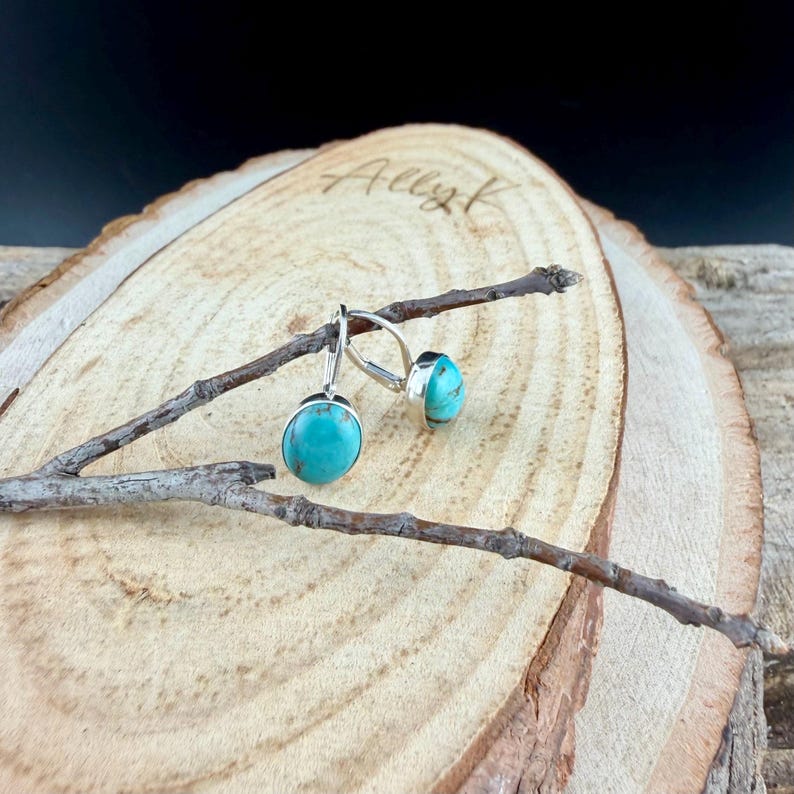 May include: A pair of turquoise earrings with a silver setting. The earrings are oval-shaped and have a bright, robin's egg blue color. They are displayed on a wooden surface with a branch.