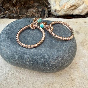 May include: A pair of rose gold hoop earrings with a twisted rope design. Each earring features a small, round turquoise stone at the top. The earrings are displayed on a gray stone, showcasing their delicate craftsmanship.