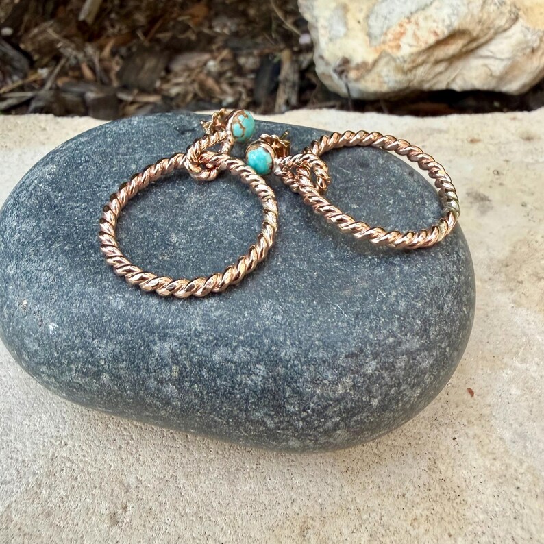 May include: A pair of rose gold hoop earrings with a twisted rope design. Each earring features a small, round turquoise stone at the top. The earrings are displayed on a gray stone, showcasing their delicate craftsmanship.