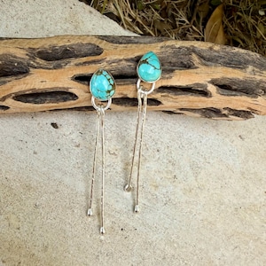 May include: A pair of dangle earrings featuring teardrop-shaped turquoise stones with brown matrix patterns. Each stone is set in a silver bezel, attached to two long, thin, textured silver wires that hang vertically from the base of the setting.
