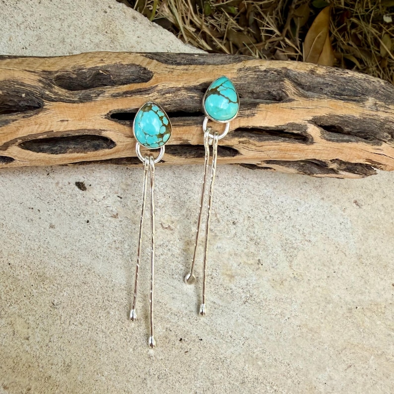 May include: A pair of dangle earrings featuring teardrop-shaped turquoise stones with brown matrix patterns. Each stone is set in a silver bezel, attached to two long, thin, textured silver wires that hang vertically from the base of the setting.