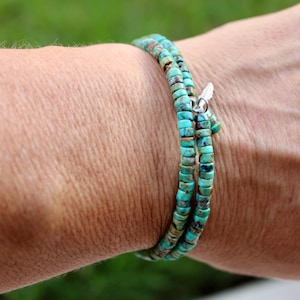 May include: A turquoise beaded wrap bracelet with a silver feather charm. The bracelet is wrapped around a wrist, showcasing the vibrant blue-green color of the stones. The bracelet appears to be made of small, flat, round beads.