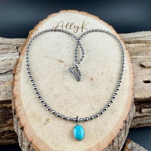 May include: A silver beaded necklace with a turquoise pendant. The pendant is an oval shape with a silver border. The necklace is displayed on a wooden surface with the text "Ally K" in the upper center.