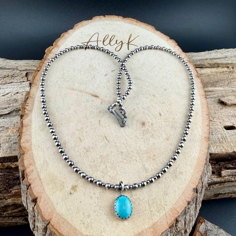 May include: A silver beaded necklace with a turquoise pendant. The pendant is an oval shape with a silver border. The necklace is displayed on a wooden surface with the text "Ally K" in the upper center.
