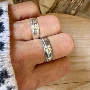 May include: Two silver bands featuring intricate engraved diamond-shaped patterns and beaded edges. The rings have a polished metallic finish and are worn on two fingers, showcasing the detailed metalwork and consistent design elements.