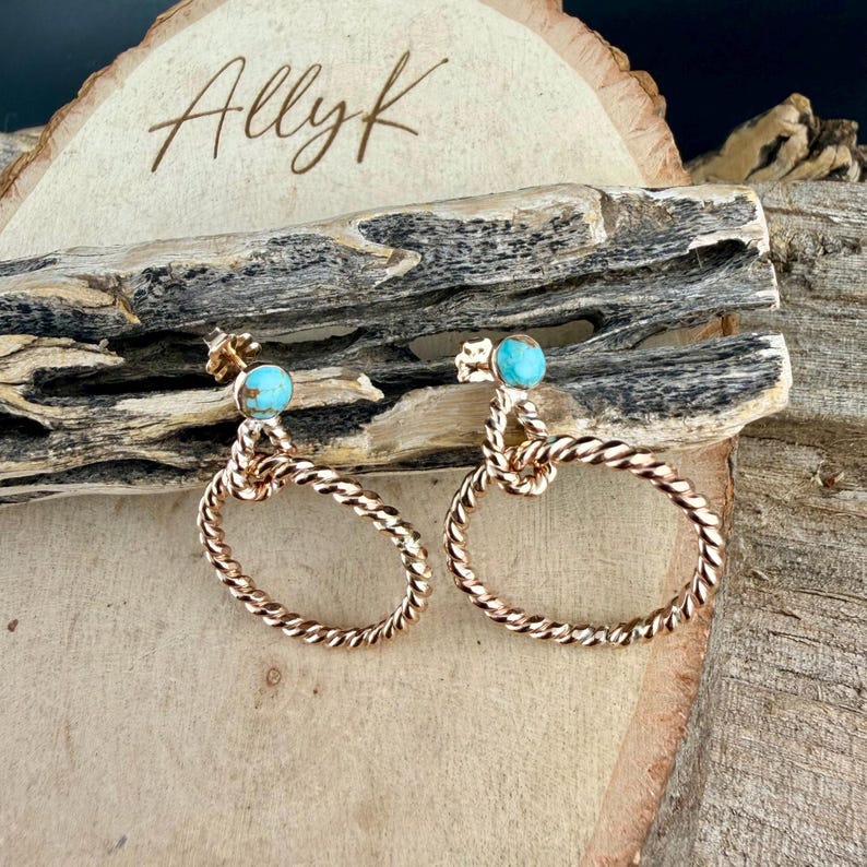 May include: Rose gold oval earrings with turquoise accents. The earrings have a twisted rope design and are displayed on a wooden surface. The earrings are a unique piece of jewelry.
