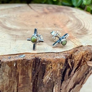 K&ouml;nnte beinhalten: Ein Paar silberfarbene Kreuz-Ohrstecker. Jeder Ohrring hat ein Kreuz-Design mit einem kleinen, runden, olivgr&uuml;nen Stein in der Mitte. Die Ohrringe werden auf einer nat&uuml;rlichen Holzoberfl&auml;che pr&auml;sentiert.