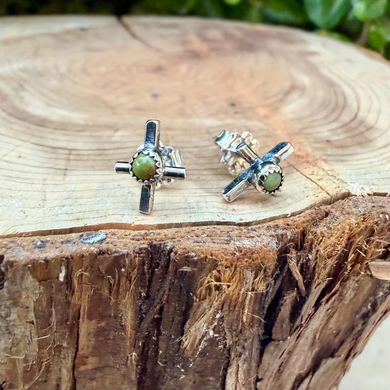 K&ouml;nnte beinhalten: Ein Paar silberfarbene Kreuz-Ohrstecker. Jeder Ohrring hat ein Kreuz-Design mit einem kleinen, runden, olivgr&uuml;nen Stein in der Mitte. Die Ohrringe werden auf einer nat&uuml;rlichen Holzoberfl&auml;che pr&auml;sentiert.