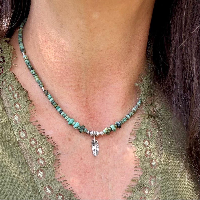 May include: A turquoise beaded necklace with a small silver feather pendant. The necklace is composed of small, irregular turquoise beads and silver spacer beads. The feather pendant hangs from a silver ring. The necklace is worn around the neck.