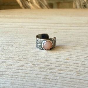 May include: A wide band ring featuring an oxidised dark silver finish with intricate etched scrollwork patterns. The centre of the band is set with a smooth, round, pale peach-coloured cabochon stone held in place by a scalloped metal bezel setting.