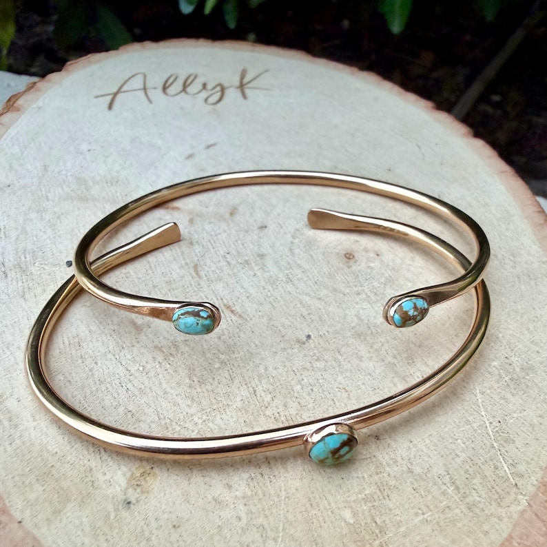 May include: Two gold-colored metal bracelets with turquoise-colored stones. The bracelets are open-ended and rest on a wooden surface. The word "Allyk" is engraved on the wood.