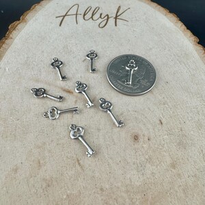 May include: Small silver-colored key charms scattered on a wooden surface, with a U.S. quarter for size comparison. The keys have intricate details and a loop at the top for attaching to jewelry or other crafts. The word "Allyk" is written in cursive at the top.