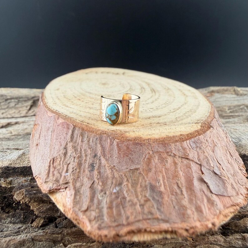 May include: A gold-colored ring with a turquoise stone. The ring has a wide band with etched details. The turquoise stone is oval-shaped and set in a bezel setting. The ring is displayed on a wooden surface.