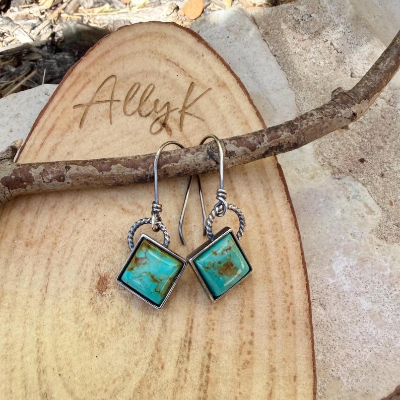 May include: A pair of silver-toned earrings with square turquoise stones. The earrings are displayed on a wooden surface with the engraved text "Allyk". The turquoise stones have brown veining.