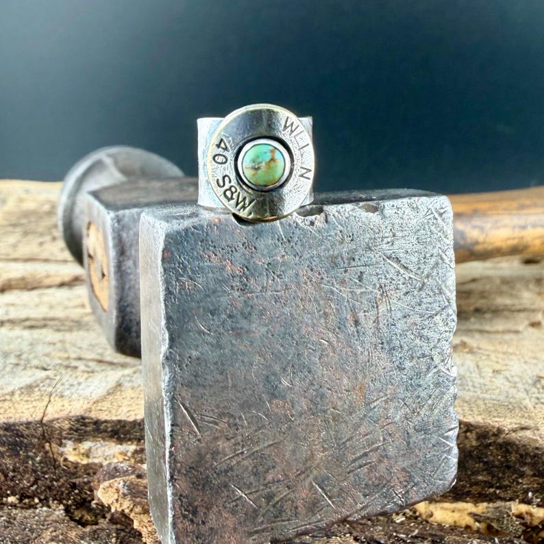 May include: A silver ring with a turquoise stone set in the centre. The ring is made from a bullet casing, with the markings "40 S&W" and "WELLN" visible. The ring is displayed on a metal hammer.