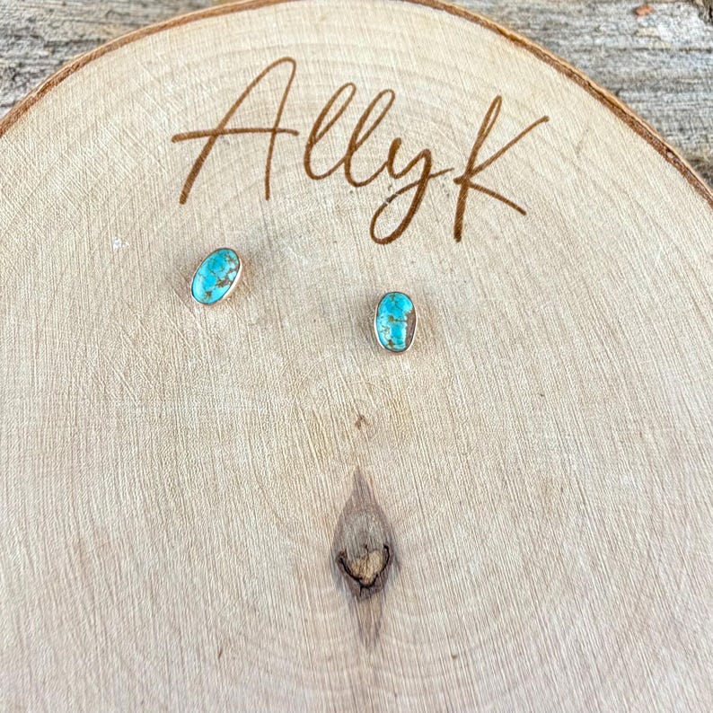 May include: A pair of turquoise-colored oval stud earrings with gold-colored metal settings. The earrings are displayed on a light brown wooden surface with the handwritten text "Allyk".