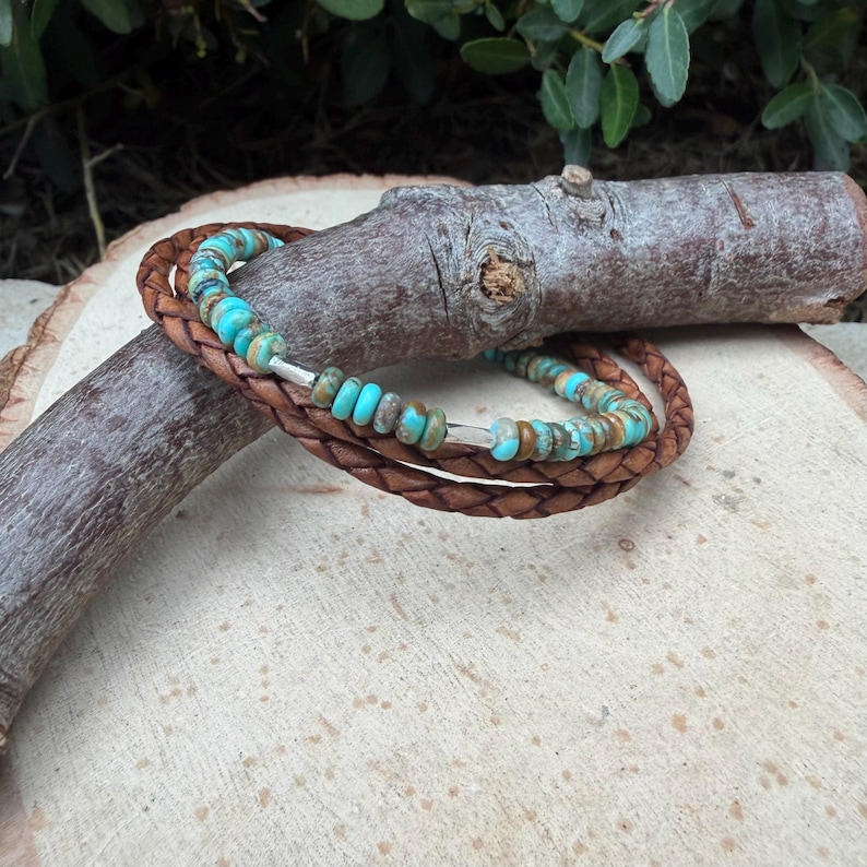 May include: A multi-strand bracelet featuring braided brown leather cords intertwined with a single strand of small, round, turquoise-colored beads with brown matrix patterns, accented by two small, smooth, silver-toned metal cylindrical spacers.