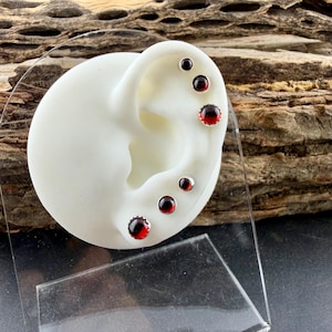 May include: A set of six silver stud earrings featuring round, deep red garnet stones. The earrings are displayed on a white ear-shaped model, with three studs along the upper cartilage and three studs along the earlobe, all set in polished silver.
