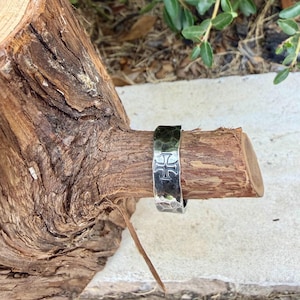 May include: A silver-toned metal ring featuring a hammered, textured surface and a prominent embossed cross symbol. The band has a wide, flat profile with a polished finish that reflects light, showcasing the intricate details of the cross design.
