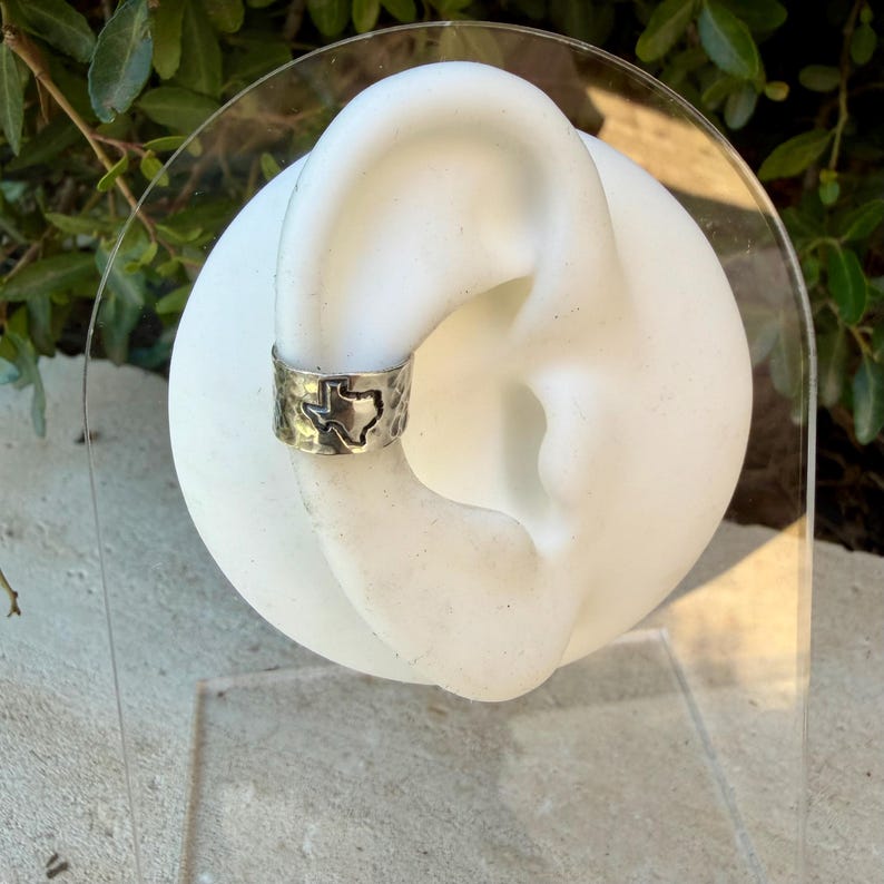 May include: A silver-toned ear cuff featuring a hammered, textured surface and a cutout silhouette of the state of Texas. The metal band is wide and wraps around the outer edge of a white, neutral-toned ear model display piece.