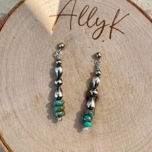 May include: A pair of dangle earrings with a silver-tone metal design. Each earring features a series of silver beads, teardrop-shaped charms, and turquoise-colored stone beads. The earrings are displayed on a wooden surface with the handwritten text "Allyk".