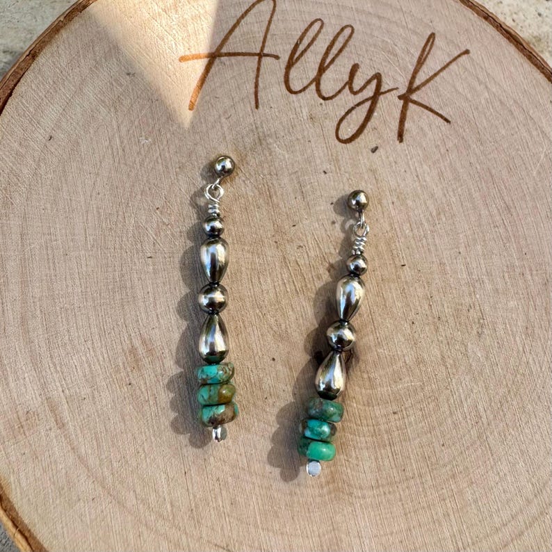 May include: A pair of dangle earrings with a silver-tone metal design. Each earring features a series of silver beads, teardrop-shaped charms, and turquoise-colored stone beads. The earrings are displayed on a wooden surface with the handwritten text "Allyk".