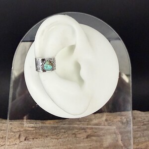 May include: A silver ear cuff with a turquoise stone. The cuff has a textured, organic design and is shown on a white ear model. The turquoise stone is a vibrant blue-green color.