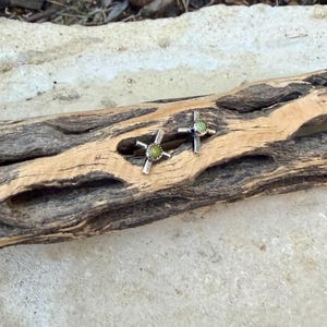 K&ouml;nnte beinhalten: Ein Paar silberfarbene Ohrstecker. Jeder Ohrring hat ein Kreuzdesign mit einem runden, hellgr&uuml;nen Stein in der Mitte. Die Ohrringe werden auf einem St&uuml;ck verwittertem Holz pr&auml;sentiert, das ihr Design und ihre Farbe hervorhebt.