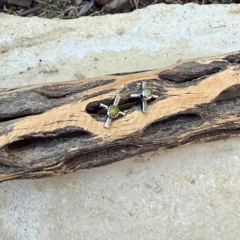K&ouml;nnte beinhalten: Ein Paar silberfarbene Ohrstecker. Jeder Ohrring hat ein Kreuzdesign mit einem runden, hellgr&uuml;nen Stein in der Mitte. Die Ohrringe werden auf einem St&uuml;ck verwittertem Holz pr&auml;sentiert, das ihr Design und ihre Farbe hervorhebt.