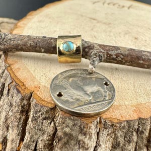 May include: A gold-colored ring with a turquoise stone set in the center. The ring is on a small branch, with a silver coin hanging below. The coin has the words "FIVE CENTS" and "UNITED STATES OF AMERICA" on it.