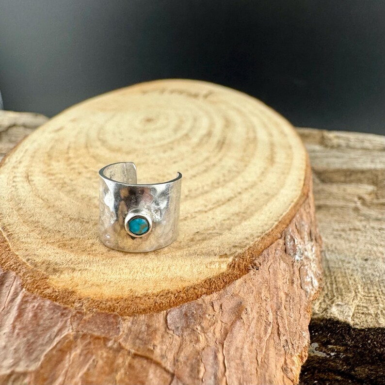 K&ouml;nnte beinhalten: Ein silberfarbener Ring mit geh&auml;mmertem Muster und einem t&uuml;rkisfarbenen Stein. Der Ring hat ein offenes Design und wird auf einer Holzoberfl&auml;che pr&auml;sentiert. Der Ring ist ein breites Band.