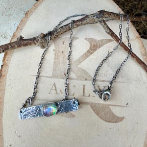 May include: A silver-toned metal necklace featuring a rectangular bar pendant with an embossed scroll pattern and a single iridescent, oval-shaped cabochon stone. The pendant is attached to a delicate, thin silver-toned chain with a lobster clasp.