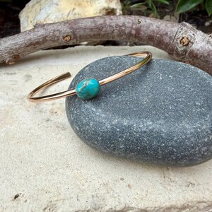May include: A gold-colored metal bracelet with a turquoise stone. The bracelet has a simple, open design with a smooth, polished finish. The oval-shaped turquoise stone is set in the center of the bracelet. The bracelet is displayed on a gray stone.
