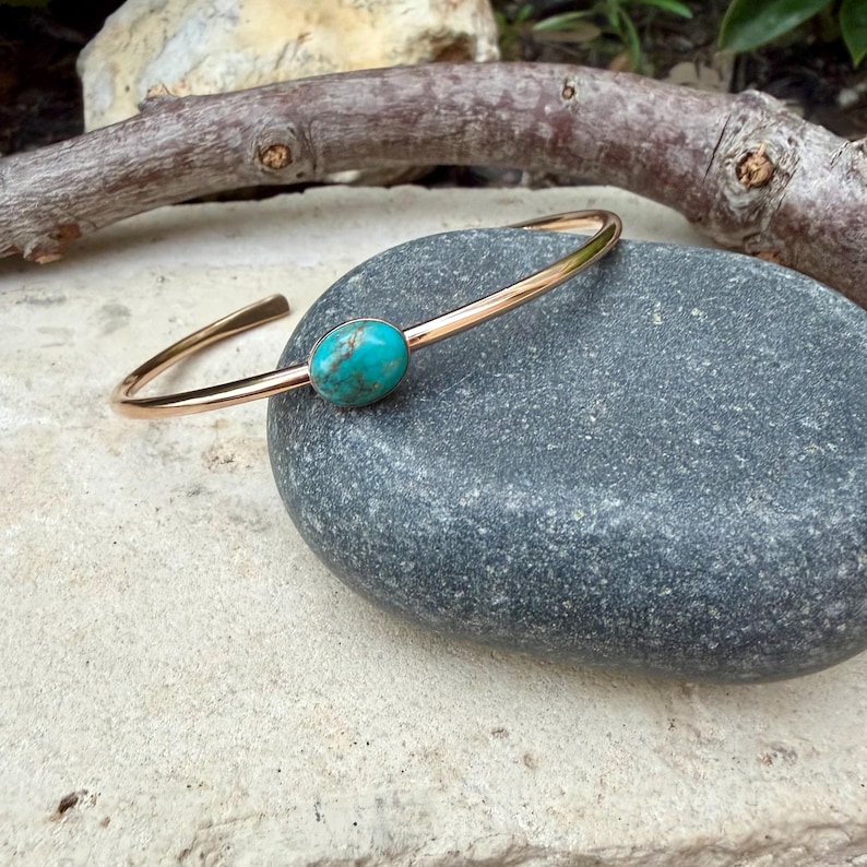 May include: A gold-colored metal bracelet with a turquoise stone. The bracelet has a simple, open design with a smooth, polished finish. The oval-shaped turquoise stone is set in the center of the bracelet. The bracelet is displayed on a gray stone.