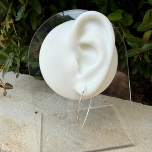 Puede incluir: Un fino alambre de pendiente de plata curvado con una peque&ntilde;a cuenta esf&eacute;rica en un extremo, exhibido en un maniqu&iacute; blanco con forma de oreja. El pendiente presenta una forma de arco alargado que cuelga del l&oacute;bulo, hecho de metal pulido.