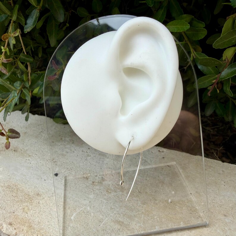 Puede incluir: Un fino alambre de pendiente de plata curvado con una peque&ntilde;a cuenta esf&eacute;rica en un extremo, exhibido en un maniqu&iacute; blanco con forma de oreja. El pendiente presenta una forma de arco alargado que cuelga del l&oacute;bulo, hecho de metal pulido.