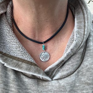 Könnte beinhalten: Eine schwarze Kordelkette mit einem silbernen Anhänger, der das Profil eines amerikanischen Ureinwohners zeigt. Die Kette hat kleine türkisfarbene Perlen. Die Kette wird über einem grauen Kapuzenpullover getragen.