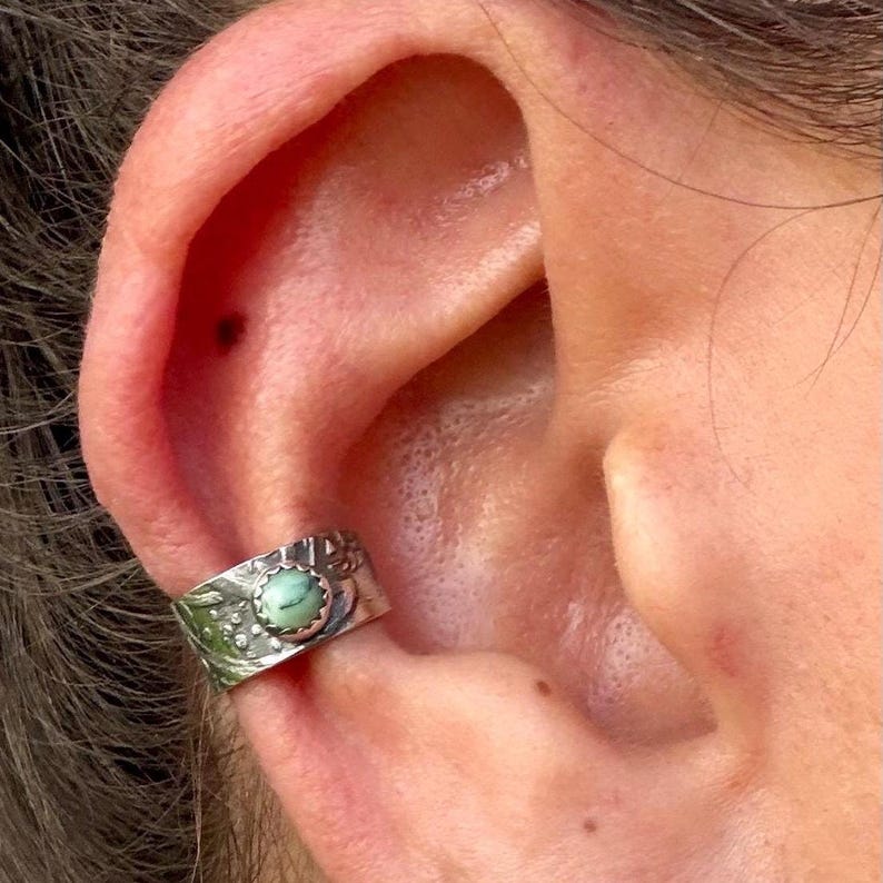 May include: Silver ear cuff with a round, light green stone. The cuff has a detailed, textured design with floral and dot patterns. The jewelry is worn on the ear, showcasing the intricate metalwork and the stone's color.