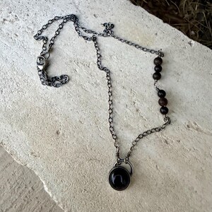 May include: A necklace featuring a dark oxidized silver chain with a circular pendant containing a smooth black stone. One side of the chain is accented with four small, round, dark brown wooden beads, and the necklace secures with a metal clasp.