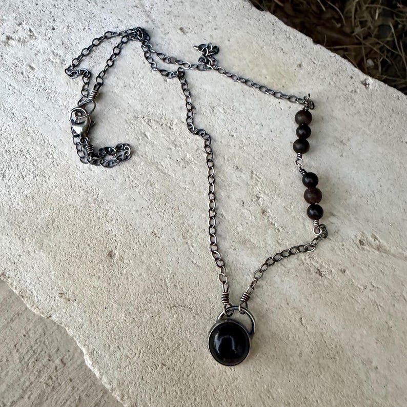 May include: A necklace featuring a dark oxidized silver chain with a circular pendant containing a smooth black stone. One side of the chain is accented with four small, round, dark brown wooden beads, and the necklace secures with a metal clasp.