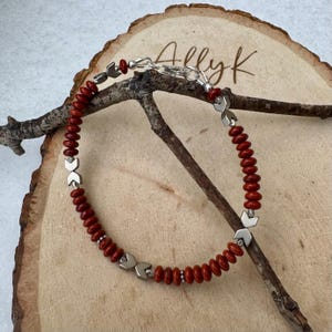 May include: A handmade bracelet featuring red beads and silver-toned metal accents. The bracelet is strung on a silver chain and rests on a wooden surface. The bracelet has a unique design with a toggle clasp.