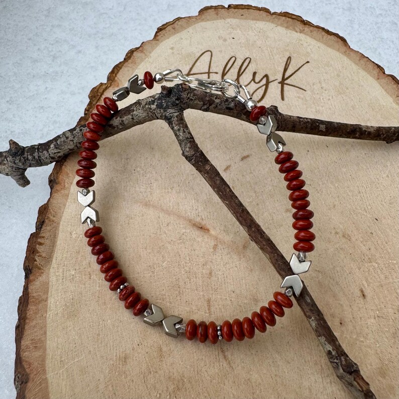 May include: A handmade bracelet featuring red beads and silver-toned metal accents. The bracelet is strung on a silver chain and rests on a wooden surface. The bracelet has a unique design with a toggle clasp.