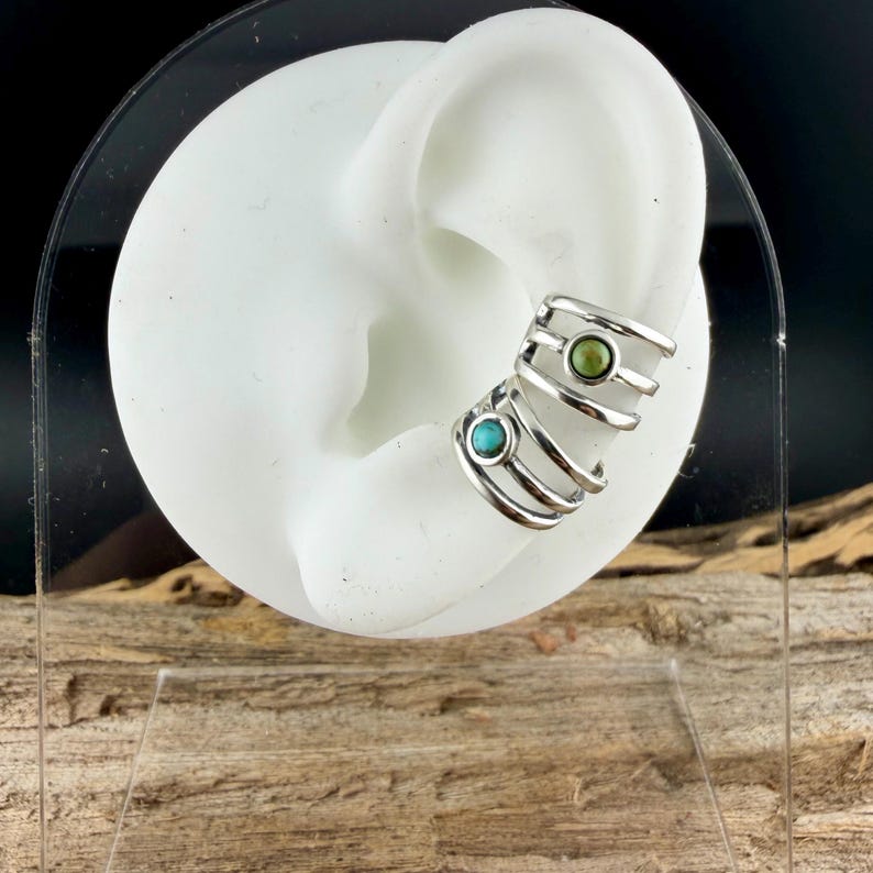 May include: Silver ear cuff with turquoise and green gemstones. The ear cuff has a layered design with multiple bands that wrap around the ear. The gemstones are set in a bezel setting. The ear cuff is displayed on a white ear model.