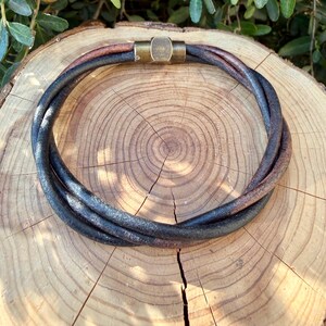 May include: A multi-strand leather necklace featuring dark brown and black twisted cords. The necklace is secured with a cylindrical antique brass magnetic clasp that connects the ends of the leather strands into a circular shape.