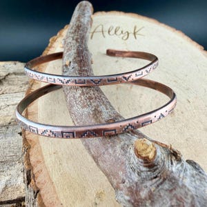 May include: Two thin copper cuff bracelets featuring a dark, etched geometric pattern of repeating squares and stepped shapes. The bracelets are open-ended and display a warm, metallic finish with oxidised details within the engraved designs.
